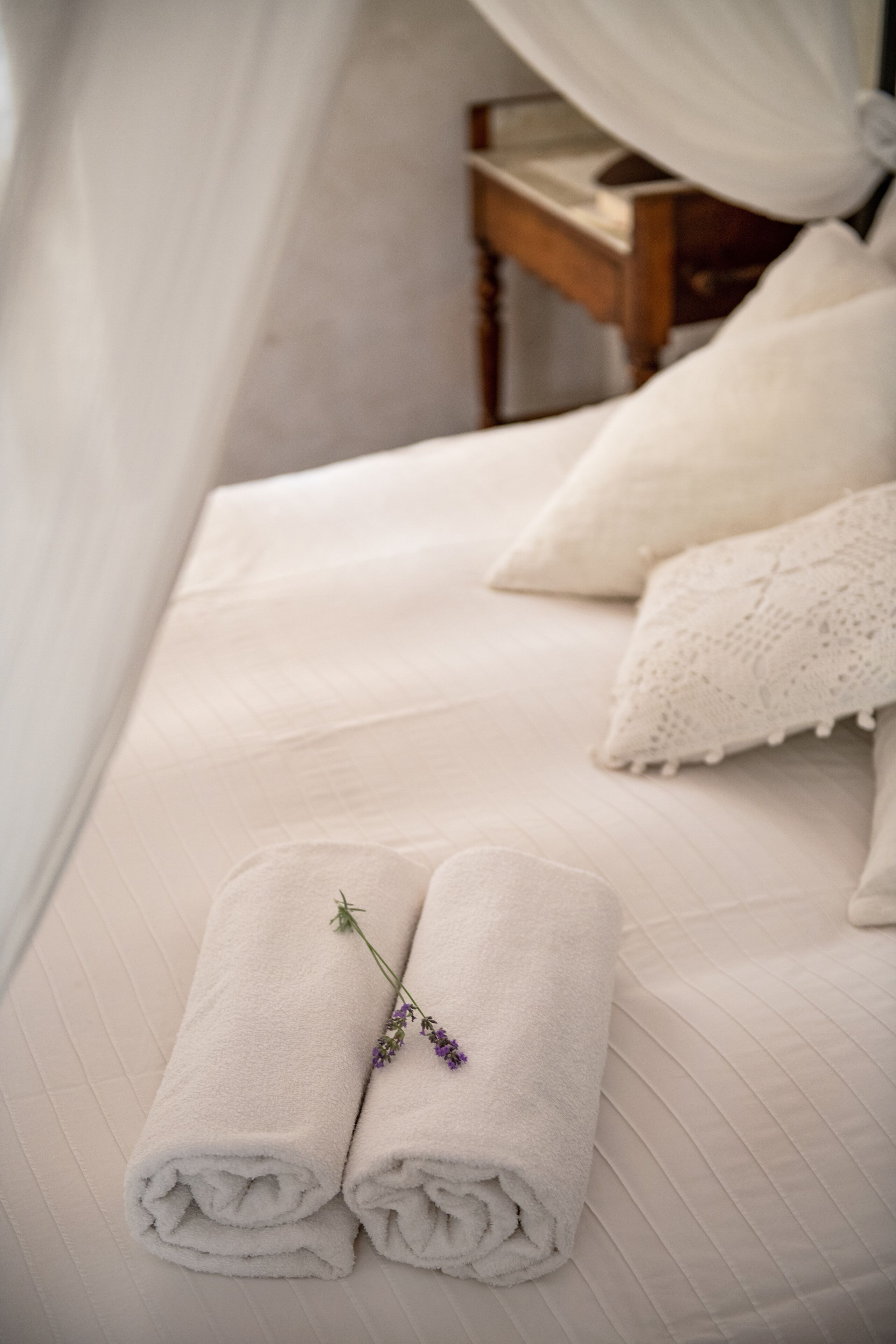 A neatly made bed with two rolled towels and a lavender sprig, a white canopy, and a wooden nightstand with a book.