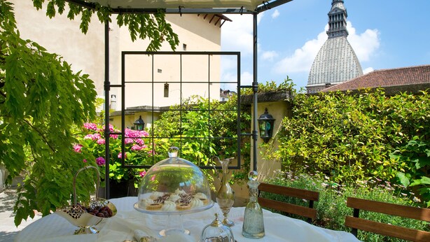 An outdoor dining area with a table set for a meal, surrounded by greenery and a view of a building with a distinctive tower. - Casa Matilde: the coziest nest in downtown Torino (Torino)