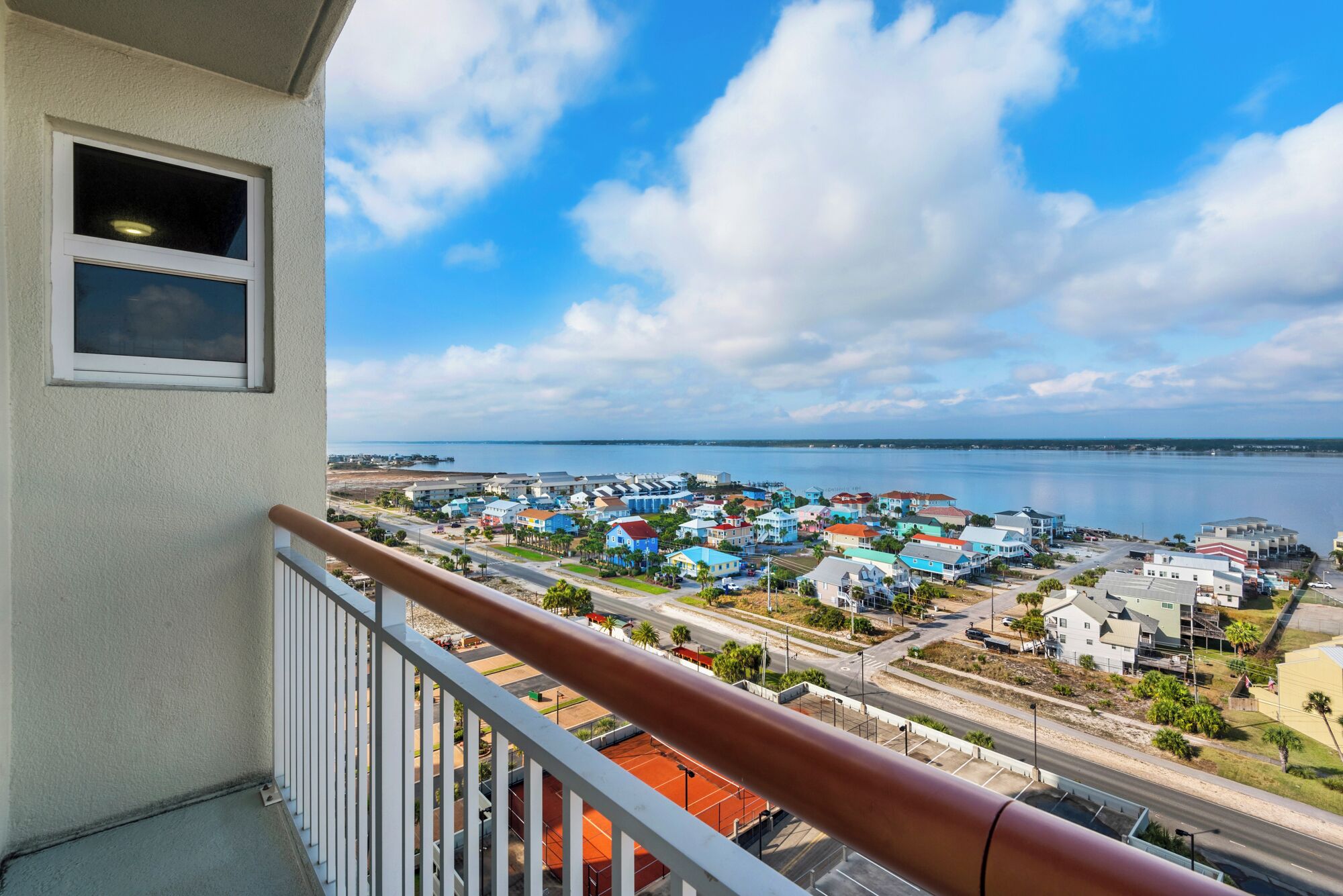 A balcony with a view of a coastal town, featuring a mix of residential and commercial buildings, a clear sky, and a calm sea.