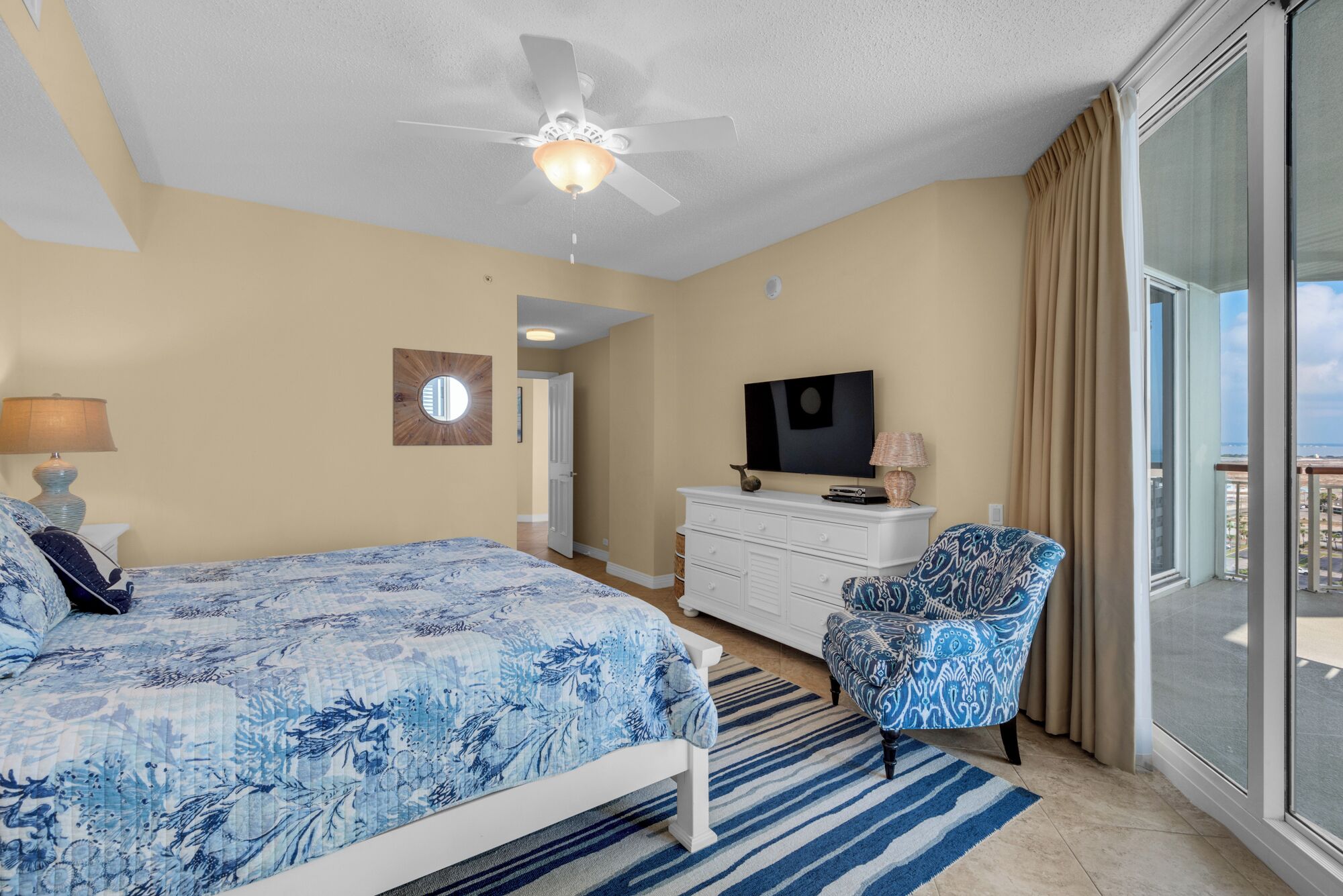 A bedroom with a bed, a chair, a dresser, a television, and a balcony with a view.