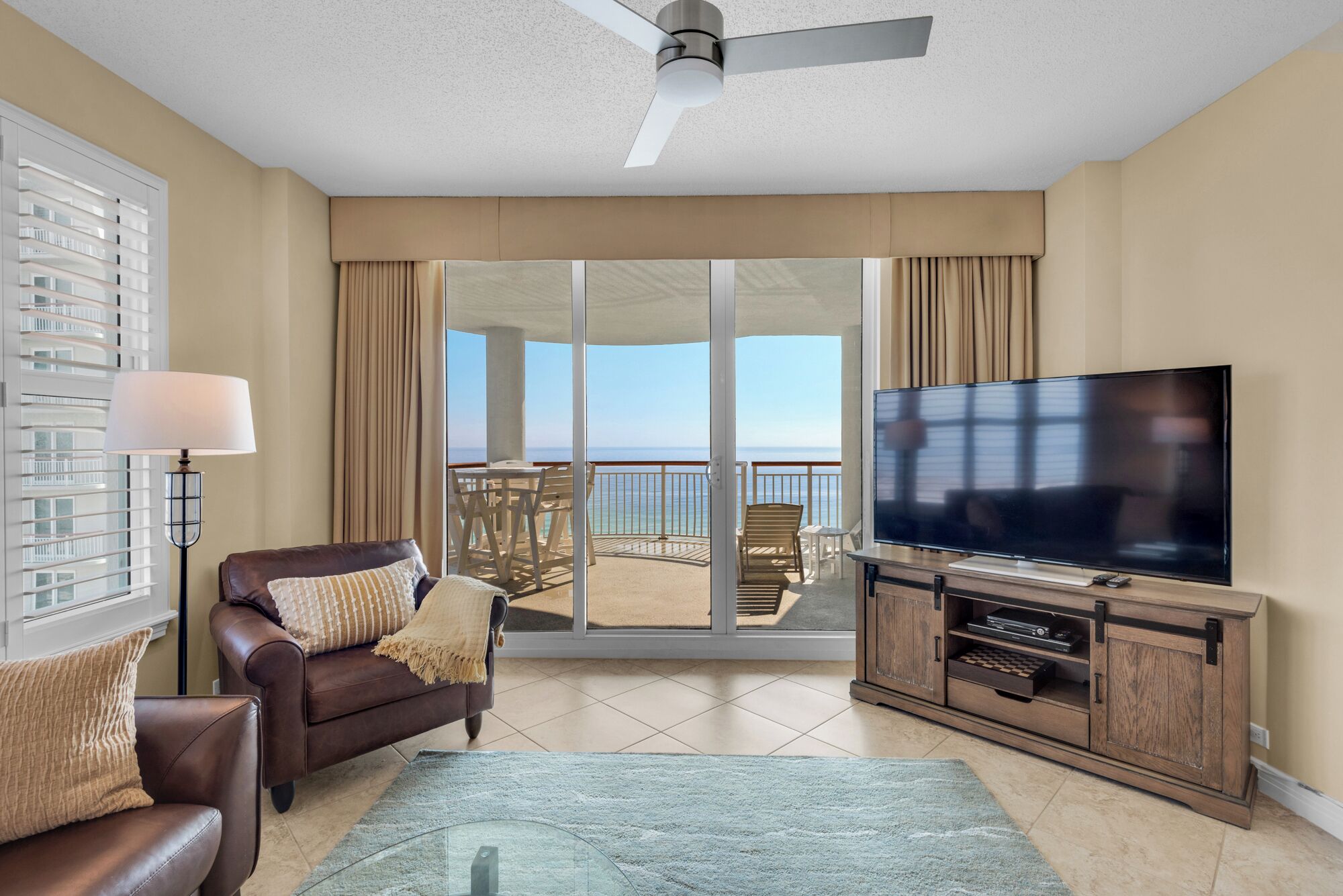 A spacious living room with a large flat-screen TV, a leather sofa, a glass coffee table, and a balcony with a view of the ocean.