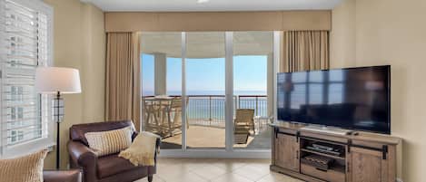 A spacious living room with a large flat-screen TV, a leather sofa, a glass coffee table, and a balcony with a view of the ocean.