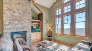 A cozy living room with a stone fireplace, leather sofas, a round coffee table, and large windows offering a view of the outdoors. - Townhome R21A-Mtn Home, Mtn View, Private Hot Tub, Indoor/Outdoor Pool & Hot Tub (Banner Elk)