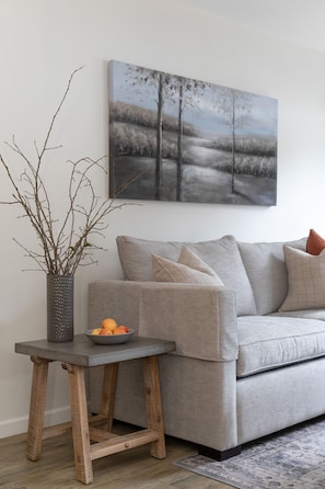 Una sala de estar con un sofá gris, una mesita de madera con un cuenco de fruta y un jarrón con ramas.