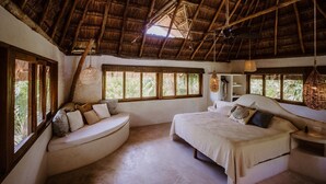 Una habitación con techo de paja, una cama, una zona de estar con asientos curvados y ventanas grandes.