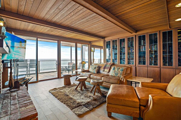 A spacious living area with a wooden ceiling, large windows, a leather sofa, and a bookshelf.
