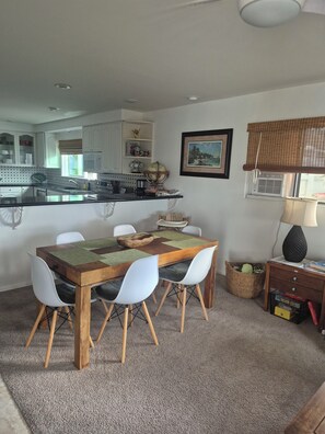 Un comedor con una mesa de madera y sillas blancas, una cocina al fondo y un cuadro en la pared.