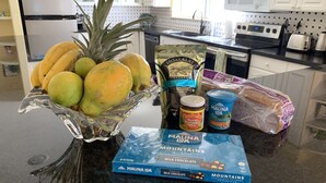 Un plan de travail de cuisine avec un panier de fruits contenant des ananas et des bananes, un pot de confiture d’ananas et de mangue, un paquet de noix de macadamia enrobées de chocolat de la marque Mauna Loa, et une miche de pain.