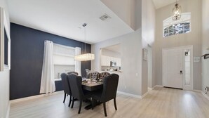 A dining area with a dark wood table and chairs, a chandelier, and a view of a kitchen in the background.