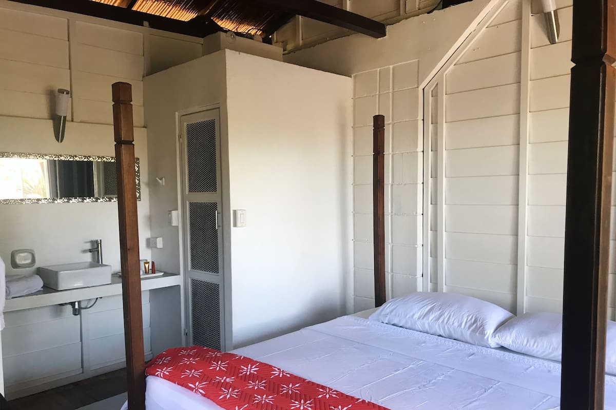 Ein Schlafzimmer mit Himmelbett, einer weißen Wand, einem kleinen Badezimmer mit Waschbecken und Spiegel sowie einem Fenster mit Jalousien.