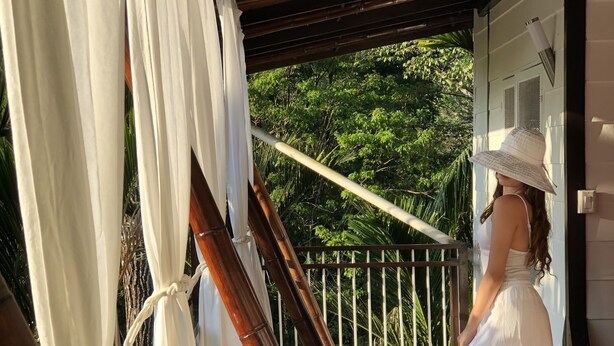 A woman in a white dress and hat standing on a wooden deck with white curtains and bamboo poles. - Ballena Dorada Hotel Boutique (Bahía Ballena)
