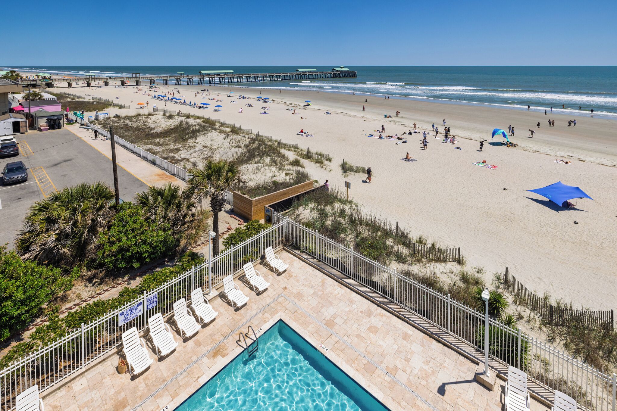 A beach view with a pool, lounge chairs, and a pier in the background.