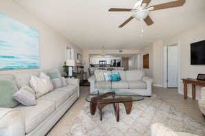 A living room with a glass coffee table, a ceiling fan, and a large painting on the wall. - Stay @ The Good Life on Folly Beach (Folly Beach)
