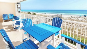 Ein Balkon mit blauen Adirondack-Stühlen und einem Tisch, mit Blick auf einen Strand mit Sonnenschirmen und Menschen.