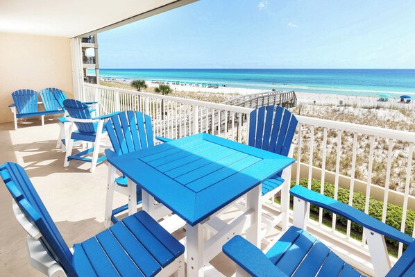 Varanda com cadeiras Adirondack azuis e uma mesa, com vista para uma praia com guarda-sóis e pessoas.