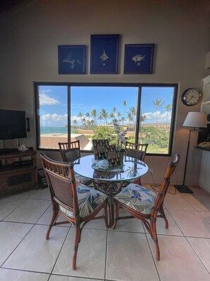 A dining area with a round glass table and wicker chairs, overlooking a beach with palm trees.