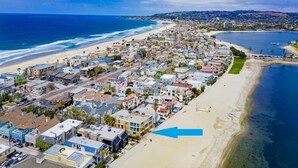 Zona residencial costera con casas a lo largo de la playa, cielo azul despejado y una flecha azul que señala una ubicación específica.