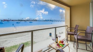 Un balcón con vistas a la playa, una mesa con frutas y una botella, y sillas de mimbre.