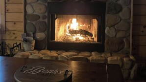 A stone fireplace with a lit fire, a wooden mantel shelf, and a coaster with 'Bari's' on it. - Enjoy our upscale, clean & well-equipped cabin in a beautiful setting. (Saugatuck)