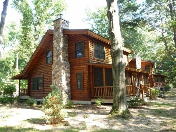 Surrounded by trees, this cabin features a stone chimney, multiple windows, and a porch with railings.