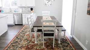 A modern kitchen with white cabinets, a stainless steel dishwasher, and a dining area with a dark wooden table and white chairs. - Beautiful Remodeled Home by Ski Resorts & Shopping (Sandy)
