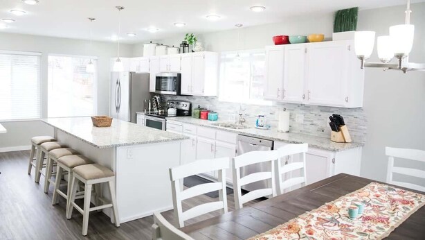 A modern kitchen with white cabinetry, a central island with stools, and a dining area with a floral rug. - Beautiful Remodeled Home by Ski Resorts & Shopping (Sandy)