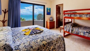 A hotel room with a bed, a bunk bed, a view of the ocean, a wooden nightstand, and a painting.