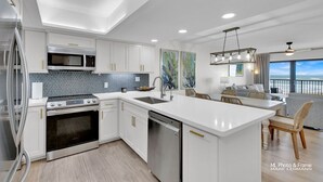 Une cuisine moderne avec des meubles blancs, un grand îlot central, des appareils électroménagers en acier inoxydable et des œuvres d’art accrochées aux murs.