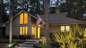 A two-story house with a prominent American flag, a well-lit entrance, and a pathway leading to the door. - Private, Immaculate 4BR/4BA Home—community pool/tennis, minutes to beach/golf (Hilton Head Island)