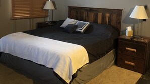 A bedroom with a bed, two bedside tables, a wall-mounted lamp, a window with blinds, and a wooden headboard. - Best Location Ski In/Out Condo @ Boyne Mountain- Walk To Village (Boyne Falls)