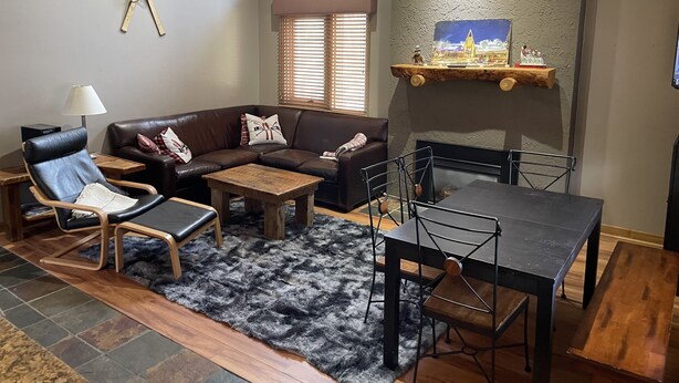 A living room with a brown leather sofa, a wooden coffee table, a black dining table with chairs, a black rug, and a fireplace with a decorative shelf above it. - Best Location Ski In/Out Condo @ Boyne Mountain- Walk To Village (Boyne Falls)