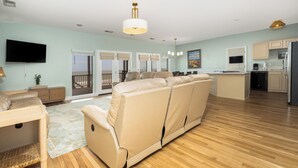 A spacious living area with a sofa, a dining table, a flat-screen TV, and a kitchen with a refrigerator. - Charleston Coast Vacations "Seascape Wonder" (Folly Beach)