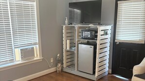 Il s’agit d’une pièce de taille modeste, comprenant un lit, une petite kitchenette équipée d’un four à micro-ondes et d’un réfrigérateur, ainsi qu’une télévision. La pièce est dotée de deux fenêtres avec stores, d’une porte et d’une affiche accrochée au mur.