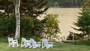 A grassy area with white Adirondack chairs facing a lake. - Oceanfront Cottage in Milbridge ~ME~ (Milbridge)