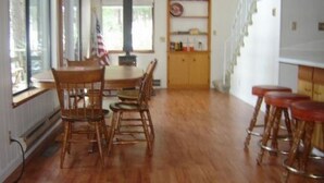 Uma área de refeições com mesa e cadeiras de madeira, um bar com banquetas, e vista para o exterior.