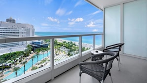 A balcony with a view of a beach and pool area, featuring wicker chairs and a table.