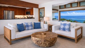 Uma sala de estar moderna com um sofá branco, almofadas estampadas em azul, uma mesa de centro de madeira e vista para o oceano através de grandes janelas.