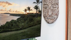 Uma televisão exibindo uma vista cênica do pôr do sol, uma máscara de madeira na parede e um armário de madeira com uma tigela decorativa e uma planta.