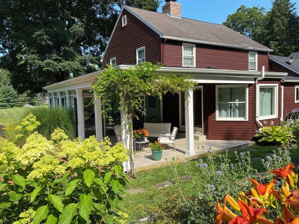 Ein Haus mit überdachter Terrasse, ein Garten mit verschiedenen Pflanzen und Blumen, und einem strahlend blauen Himmel.