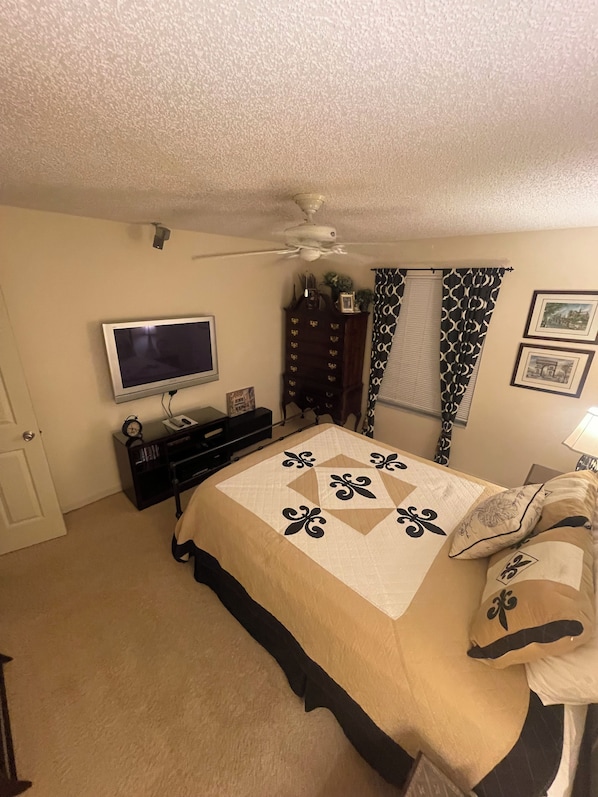 Un dormitorio con cama, televisor, ventilador de techo, cómoda y ventana con cortinas estampadas.