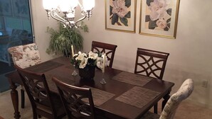 A dining area with a wooden table, six chairs, a chandelier, and two framed floral paintings on the wall.