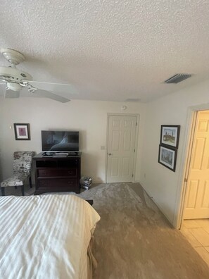 Un dormitorio con cama, televisor sobre un mueble, silla, puerta y ventilador de techo.