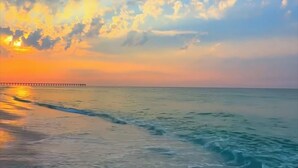 A beach with a pier extending into the water, waves gently lapping at the shore, and a sunset in the background.