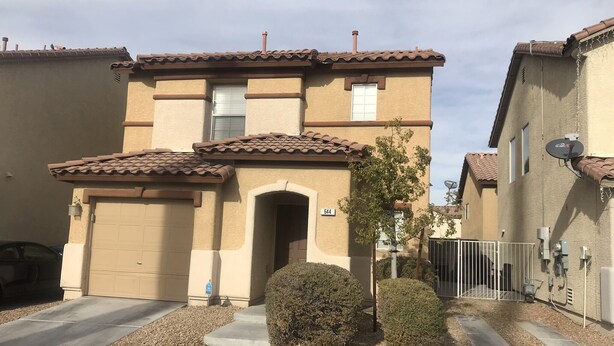 Un immeuble résidentiel à la façade beige, doté d’un portail de garage et d’un petit jardin devant avec un arbre et des arbustes.