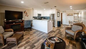 A modern living room with a large flat-screen TV, wicker furniture, and a kitchen area with white cabinets and a central island.
