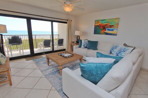 A living room with a white sofa, blue cushions, a woven coffee table, and a painting of boats on the wall. - Shore to Please - Condo 402: A cozy oceanfront  2-bedroom retreat in South Padre Island. (South Padre Island)