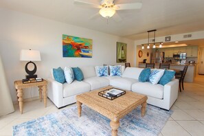 A living room with a sectional sofa, a coffee table, and a painting on the wall. - Shore to Please - Condo 402: A cozy oceanfront  2-bedroom retreat in South Padre Island. (South Padre Island)