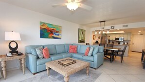 A living room with a blue sofa, a coffee table, and a painting of boats. - Shore to Please - Condo 402: A cozy oceanfront  2-bedroom retreat in South Padre Island. (South Padre Island)