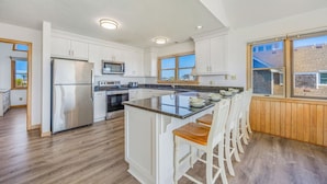 A modern kitchen with white cabinets, stainless steel appliances, a central island with bar stools, and large windows offering views.