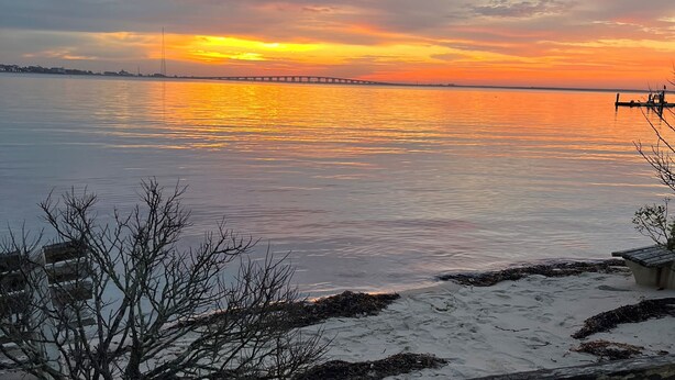 A sunset over a calm body of water with a distant bridge and a sandy shoreline with seaweed. - Relax and enjoy this private location in Hampton Bays (Hampton Bays)